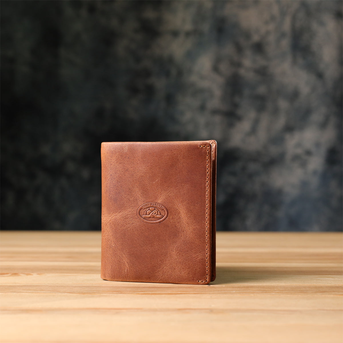 Tony Perotti Arno 1165 whisky brown leather bifold wallet standing on a wooden surface against a dark textured background