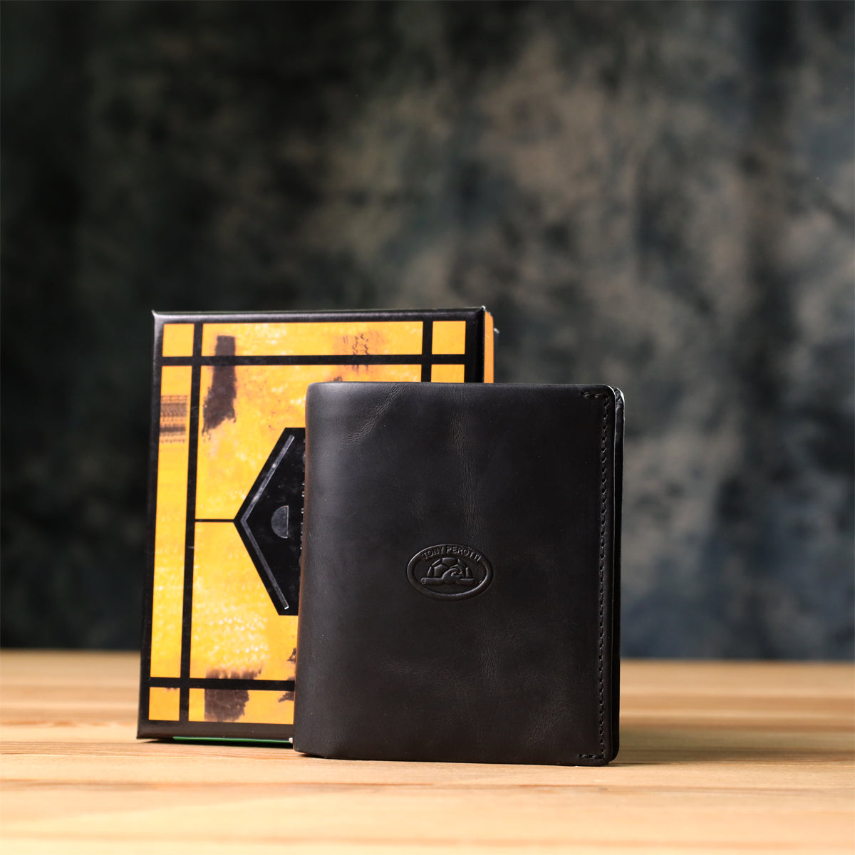 Tony Perotti Arno 1165 nero black leather bifold wallet standing next to its marbled orange and black printed gift box on a wooden surface