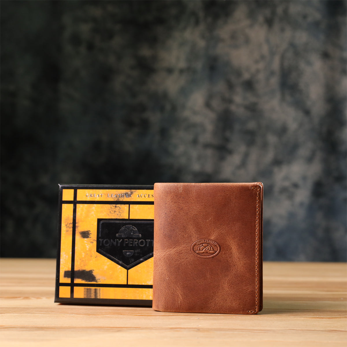 Tony Perotti Arno 1165 whisky brown leather bifold wallet standing next to its marbled orange and black printed gift box on a wooden surface