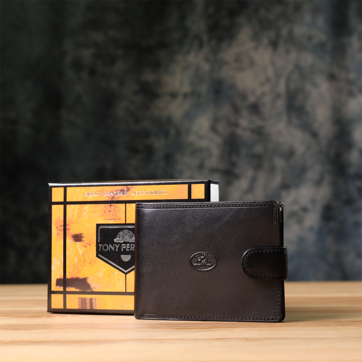 Tony Perotti Italico 396A-Q nero black leather bifold wallet standing next to its marbled orange and black printed gift box on a wooden surface