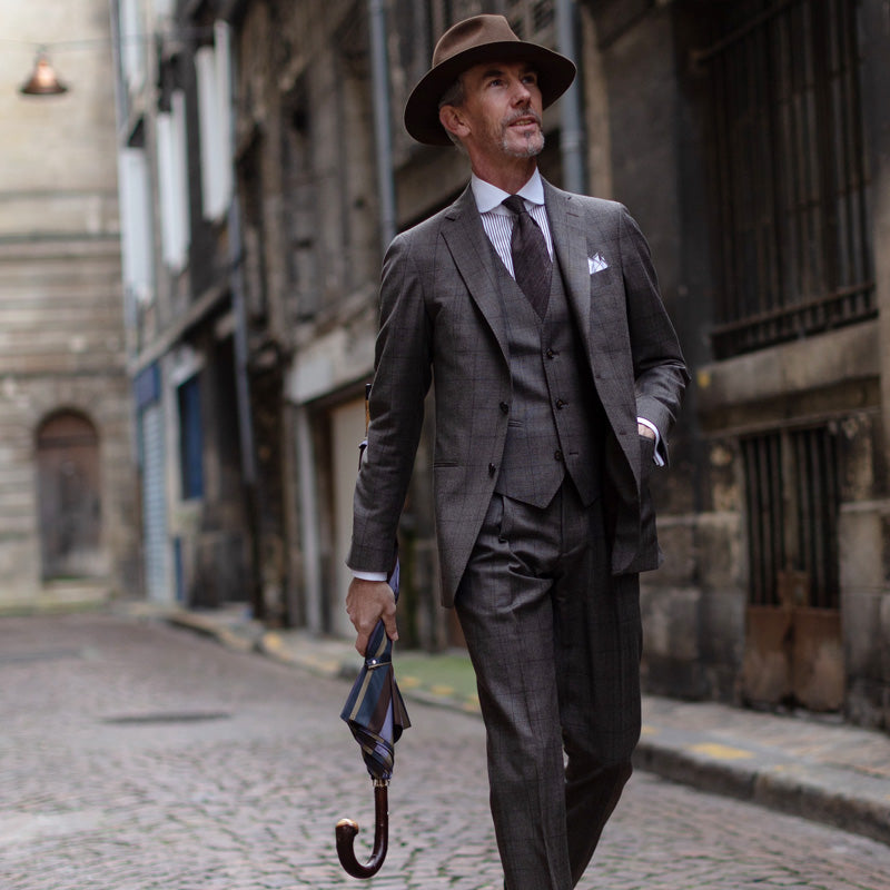 Man in a grey three-piece suit walking down a cobbled street with the furled striped umbrella