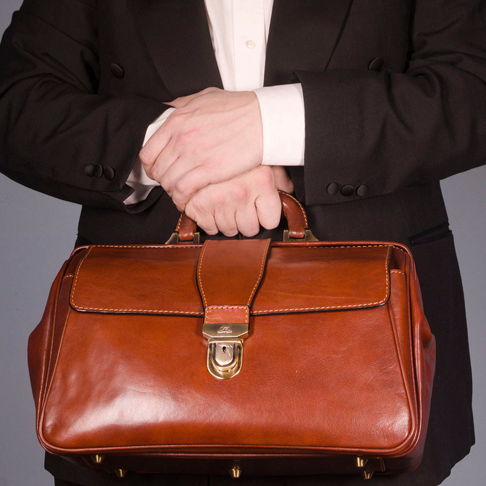 A cognac leather briefcase held by a person wearing a black blazer and white dress shirt, shown from the chest