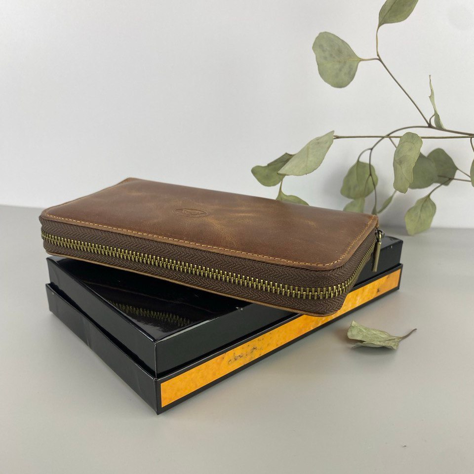 Brown leather wallet with gold zipper on a black and yellow box, with green leaves in the background.