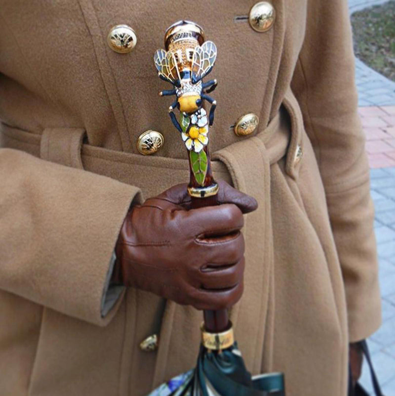Person holding a decorative umbrella handle with a bee design, wearing a brown coat and gloves.