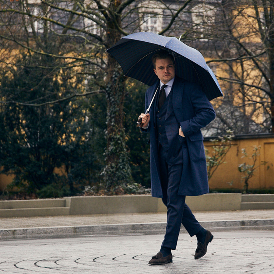 Man in a navy suit and coat walking under the open umbrella