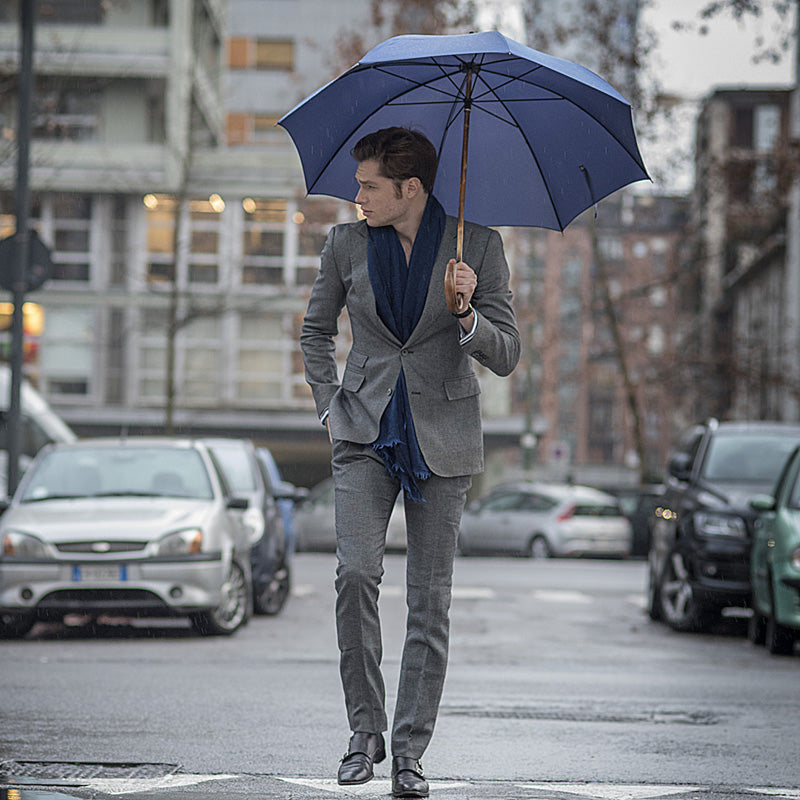 Man in a grey suit walking down a city street under the open blue umbrella